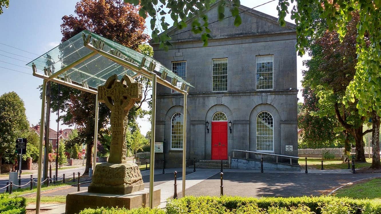 A two storey grey stone building with a red door and a Celtic cross in front