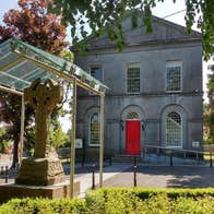 A two storey grey stone building with a red door and a Celtic cross in front