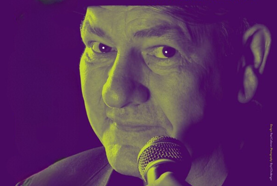 Close up head shot of man holding mic up close to face looking sideways from his left at the camera, face lit by lime green light against a dark background.