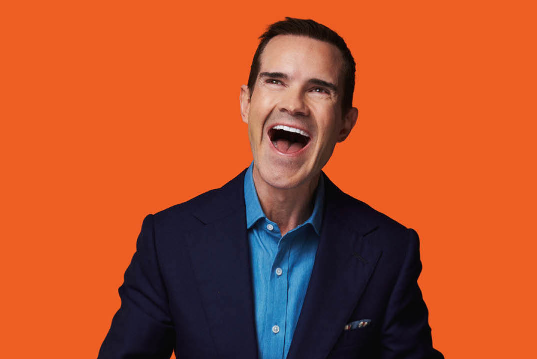 A man in dark blue jacket and shirt is laughing out loud, against orange background.