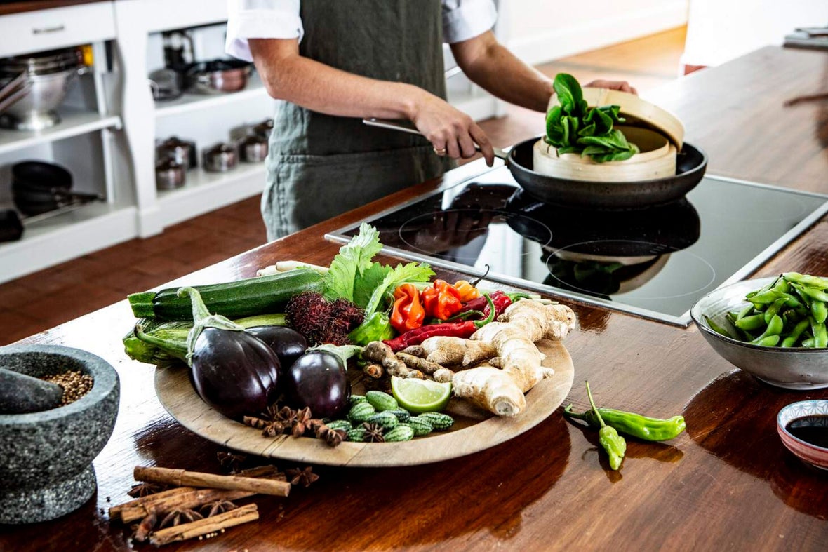 A chef preparing food over a stove with many different vegetables on his brown wooden counter top.