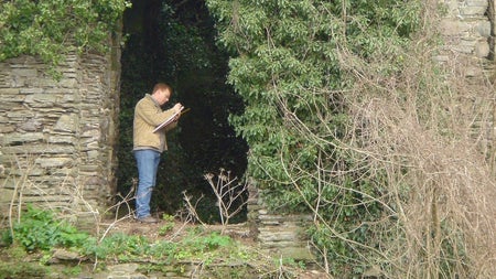 A person standing writing on a notebook within a dark opening in a historic stone structure