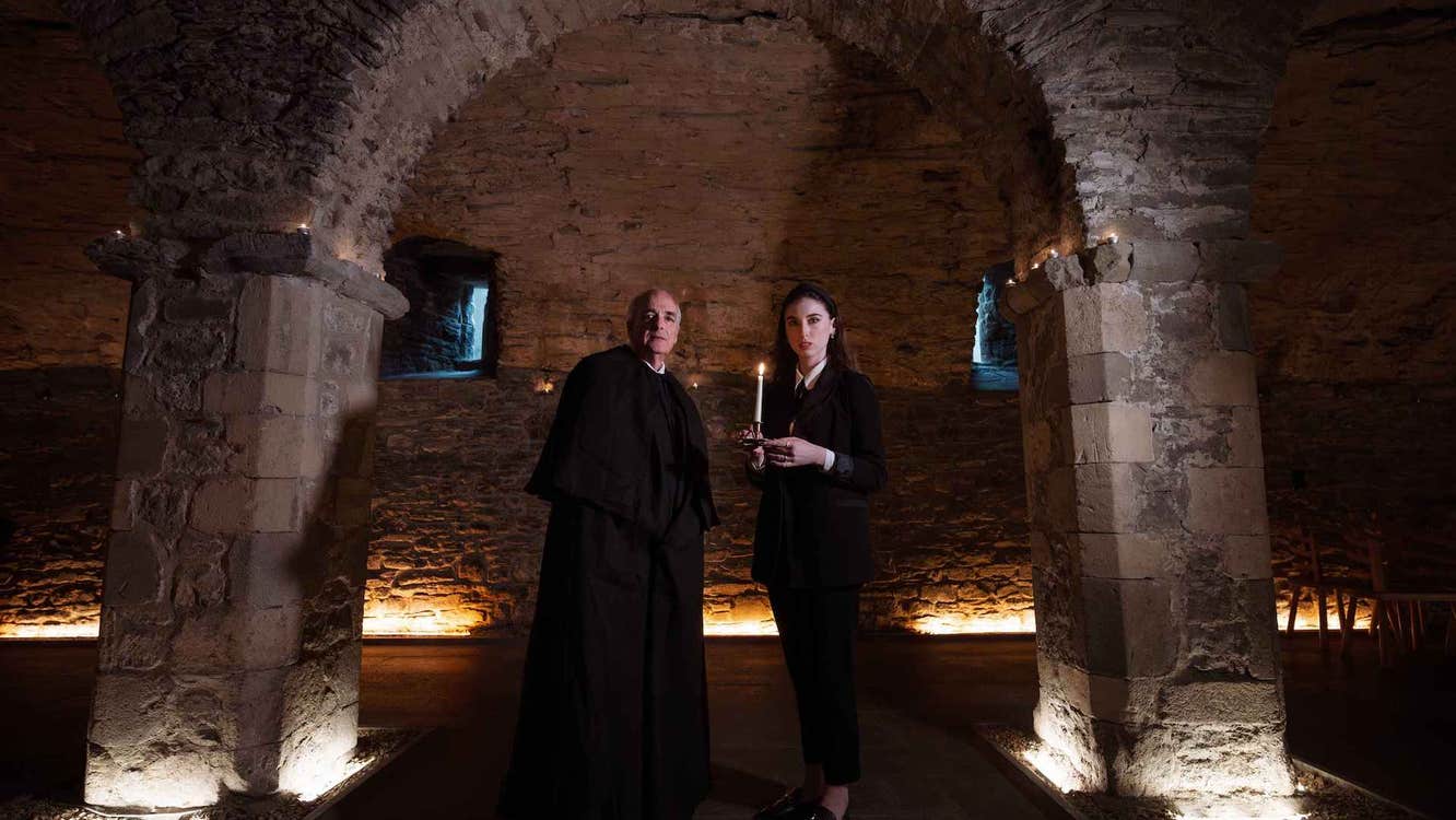 A darkly lit stone room, 2 people dressed in black are holding candles standing under a stone arch.