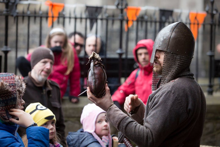 A group of adults and children are looking at a man dressed in Viking attire holding up a small, brown object in one hand.