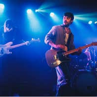 Cian Godfrey of Somebody’s Child plays guitar on stage, with drums visible behind him and atmospheric lights creating a vibrant backdrop.