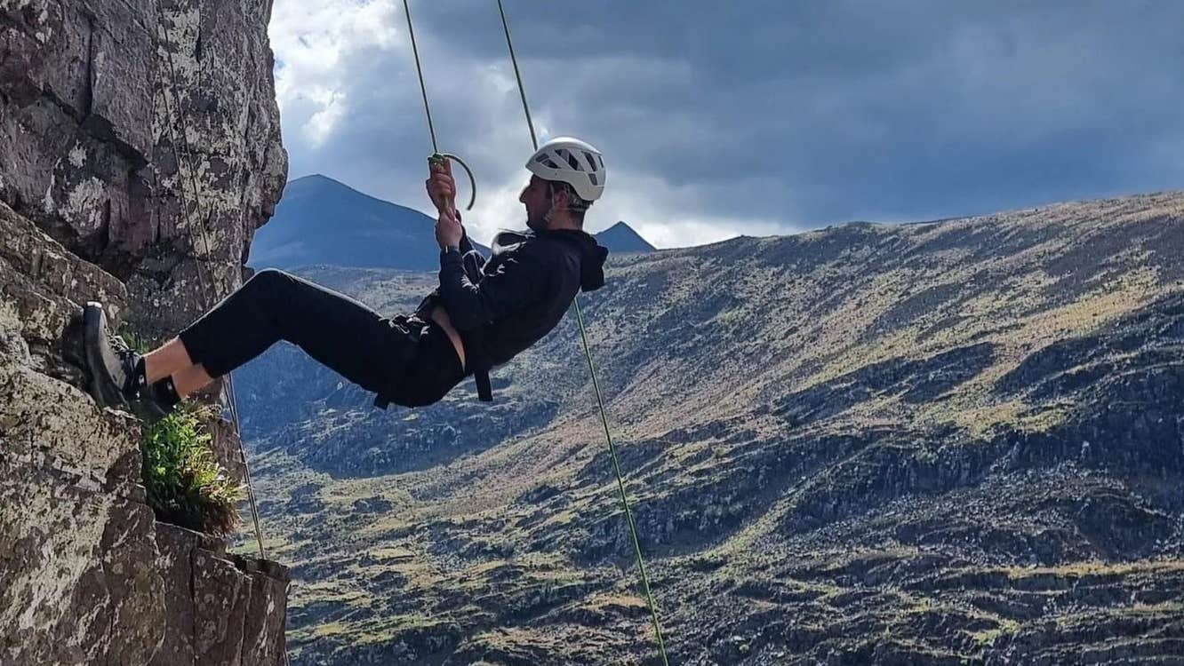 A man abseiling down a rock face with dramatic mountain and sky view in the background