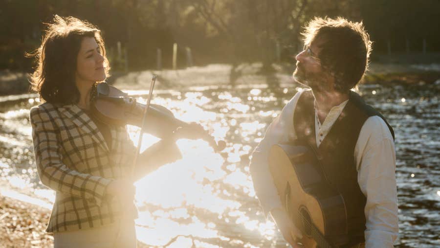 A woman playing a fiddle looking at a man playing a guitar, both standing outside with water and trees in the background.