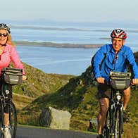 A man and woman cycling up a hill with a view of the sea visible behind them