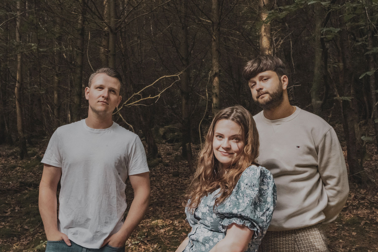 2 men and a woman are standing in front of dense woods, smiling gently.