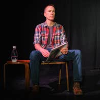a man in checked shirt seated on a chair on stage