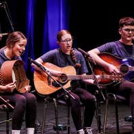 Young adults seated, playing musical instruments, singing as part of a larger group on a stage