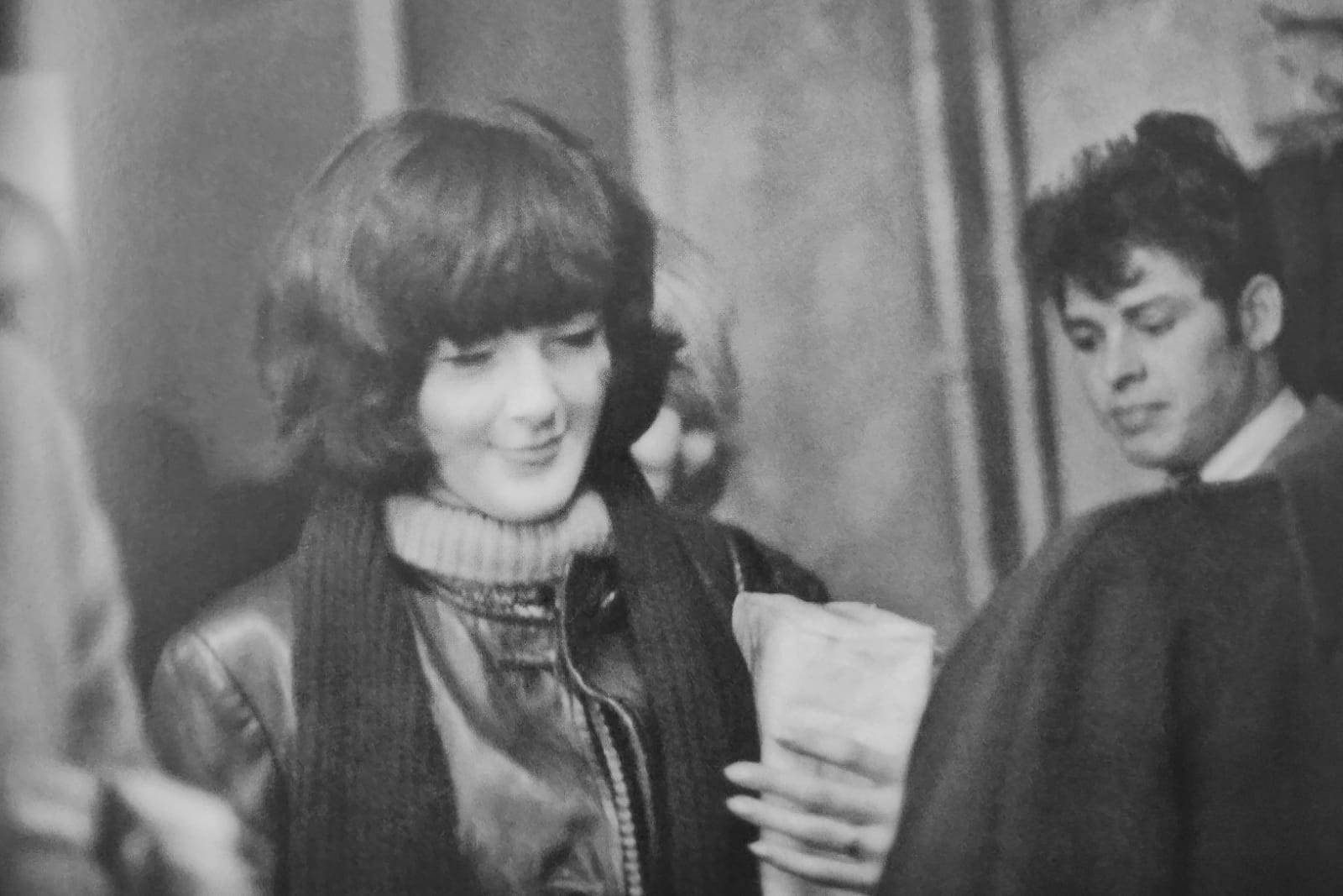 Old black and white photo of a woman in leather jacket and scarf, looking downwards, smiling with face of a man in the background.