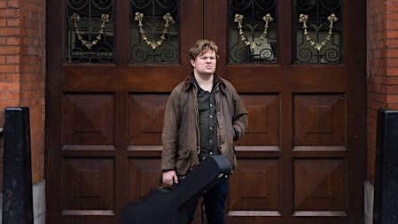 A man holding a guitar case is standing in front of large, dark wooden doors.