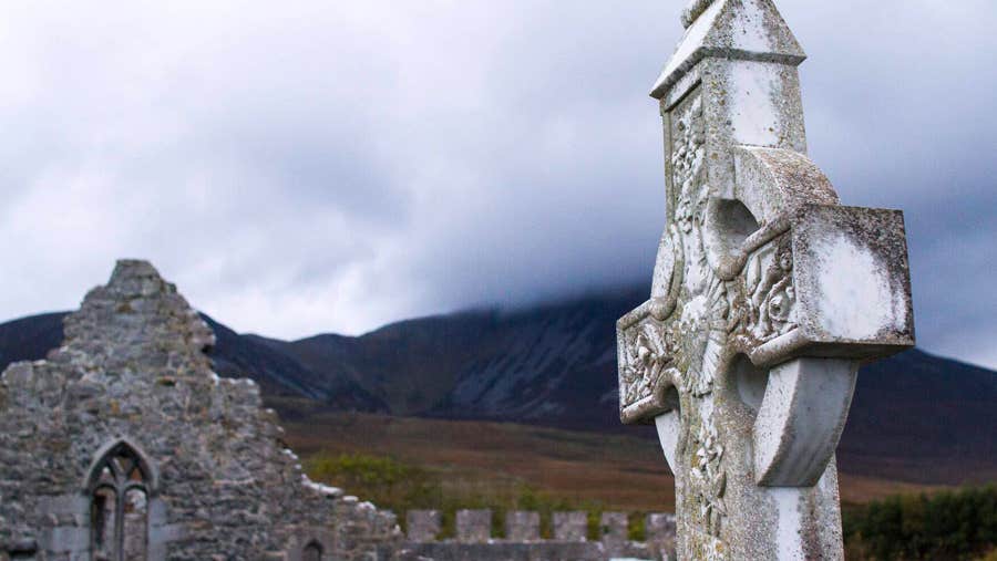The ruins of an abbey in the background with a large high cross in the foreground.