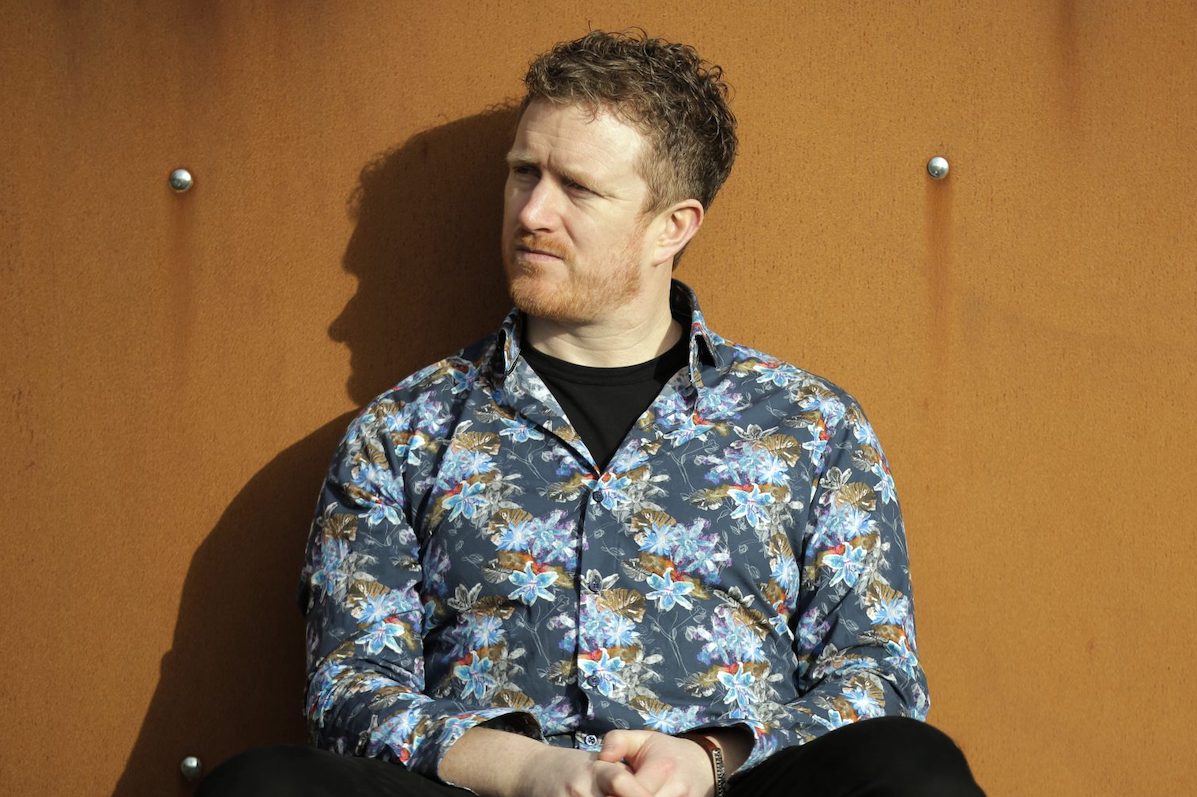 A man sat up against a dark orange wall outdoors, he is wearing a mixed blue floral shirt and dark trousers, looking away to his right
