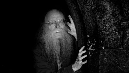 An older man in glasses with long busy beard, peering around a tree trunk in the dark.
