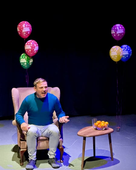 A man sits in an arm chair with Birthday balloons in the background