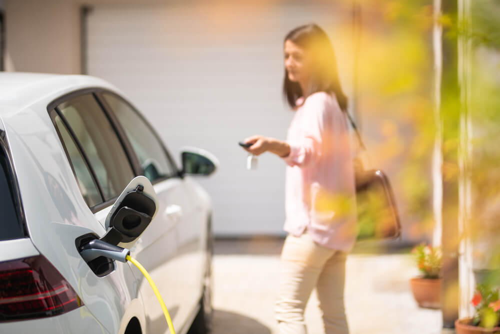 A woman locking her electric car which is parked in front of a garage with a charging cable