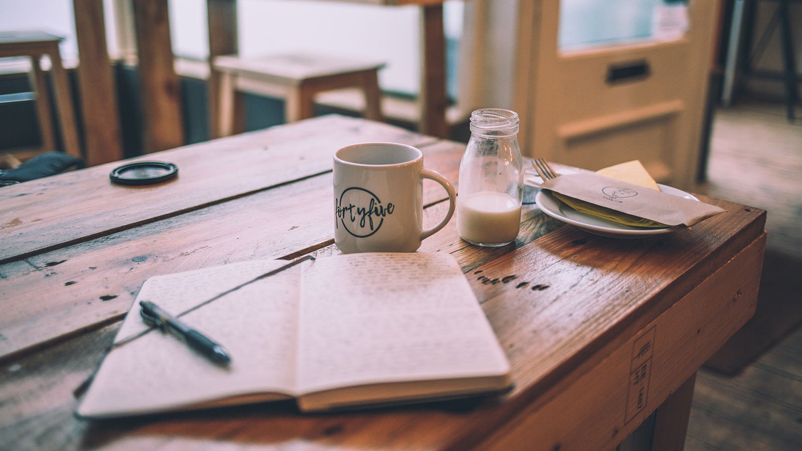 Writing in an open note pad on a wooden table in a cafe with a cup