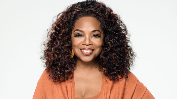 A photograph of Oprah Winfrey in an orange top against a white background.