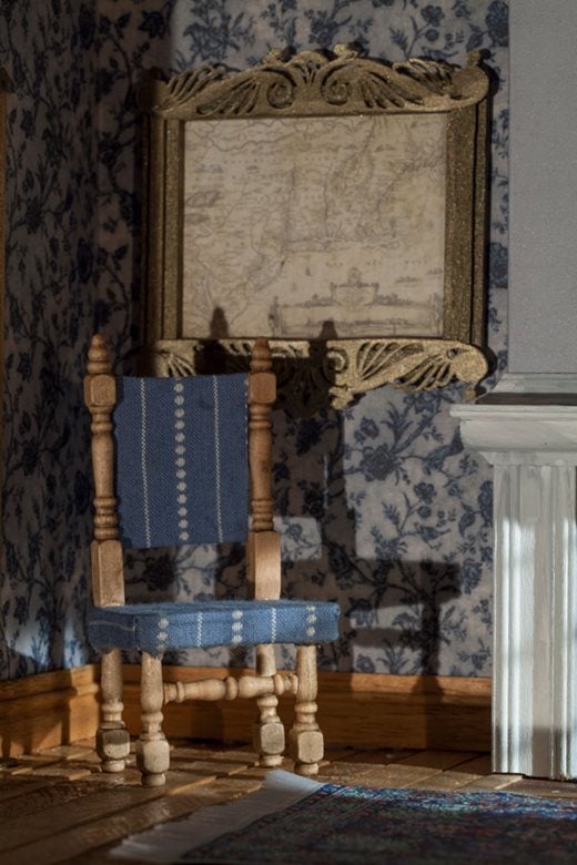 Close up image of the tiny blue chair, behind on the wall a tiny framed map is visible