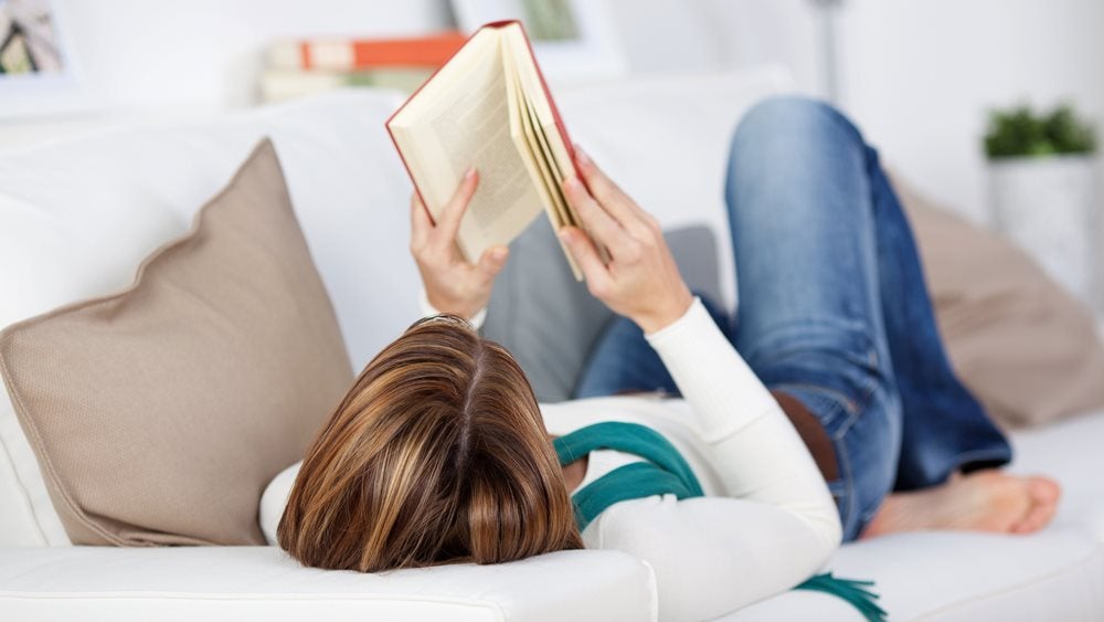 A photo of a woman lying on her sofa, reading a book