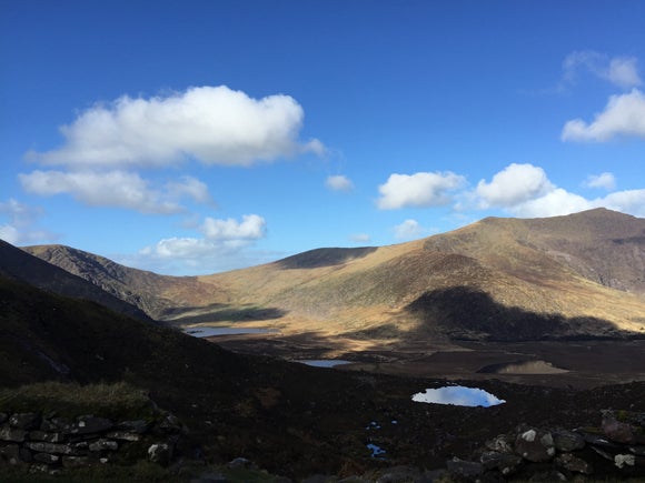 A landscape scene of mountains and a valley with small lakes within in, and clouds above in a bright blue sky casting shadows on the valley beneath 