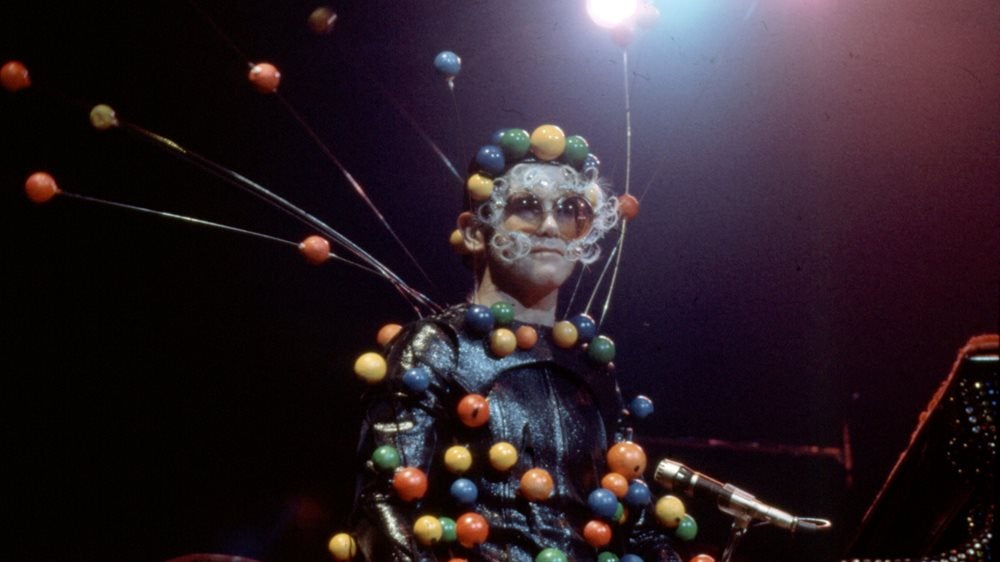 Elton John perfoming with a black suit covered in multicoloured balls and a headdress made of balls