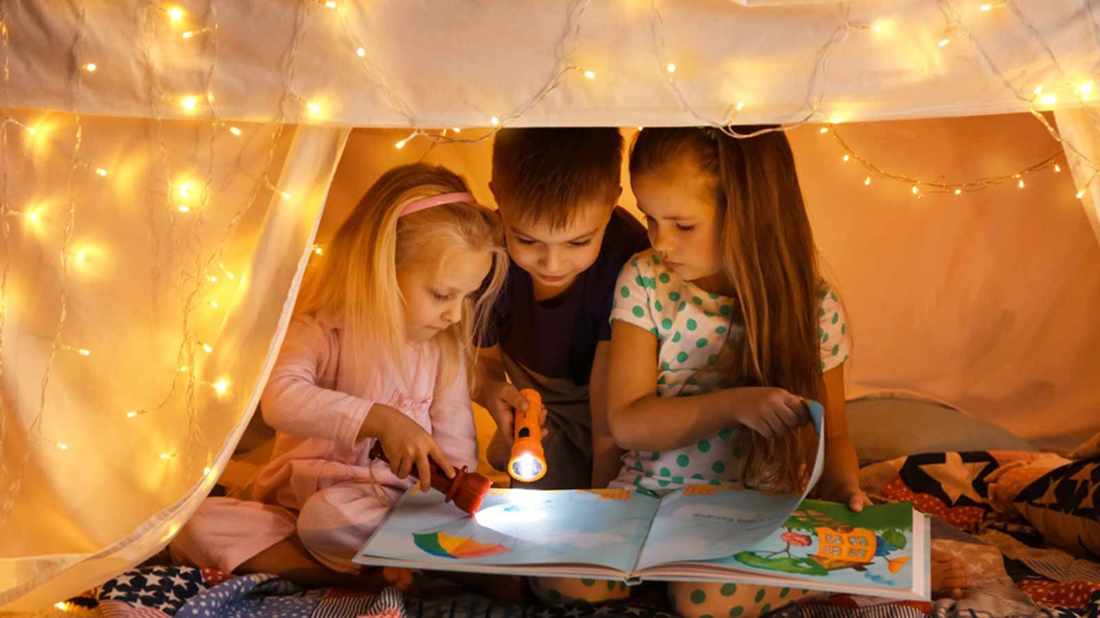 Little boy and two little girls reading a book by torchlight in a fairy light-covered tent