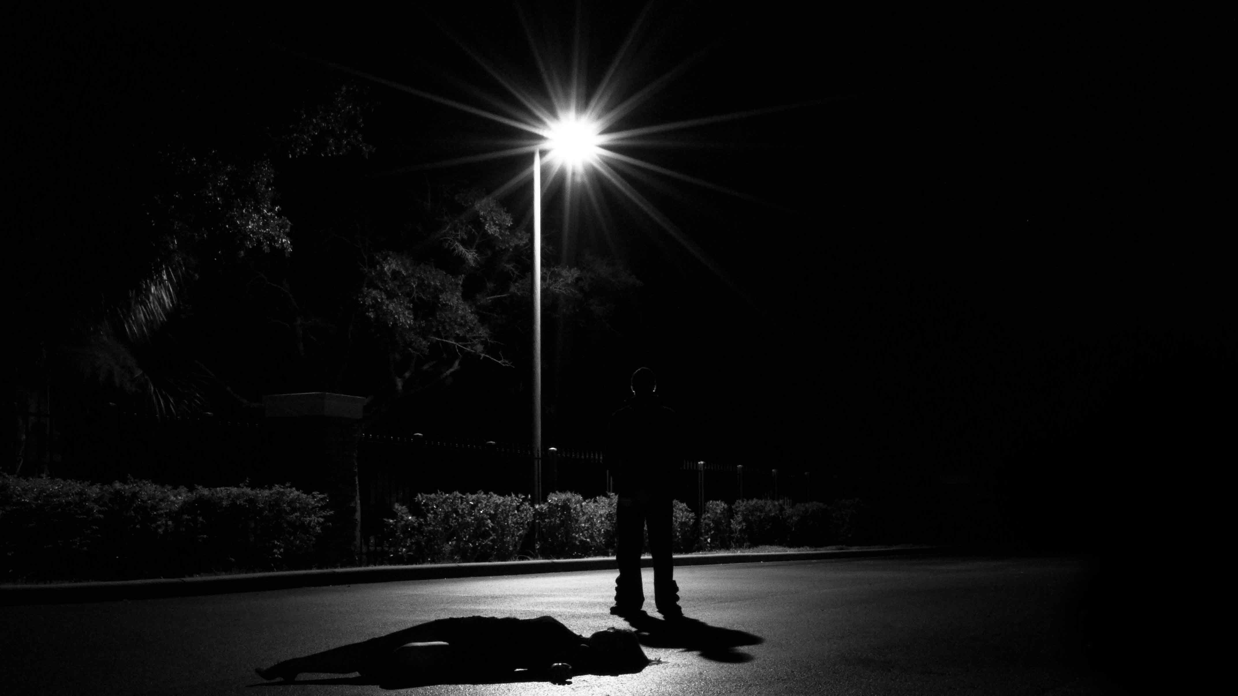Dark figure standing beneath a street light over an unconscious figure