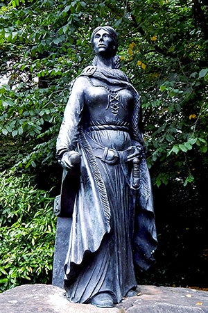 A photograph of a statue of Grainne Mhaol Ni Mhaille. She stands, looking out to see, in a shawl, her hand resting on her belt. She has a sword.