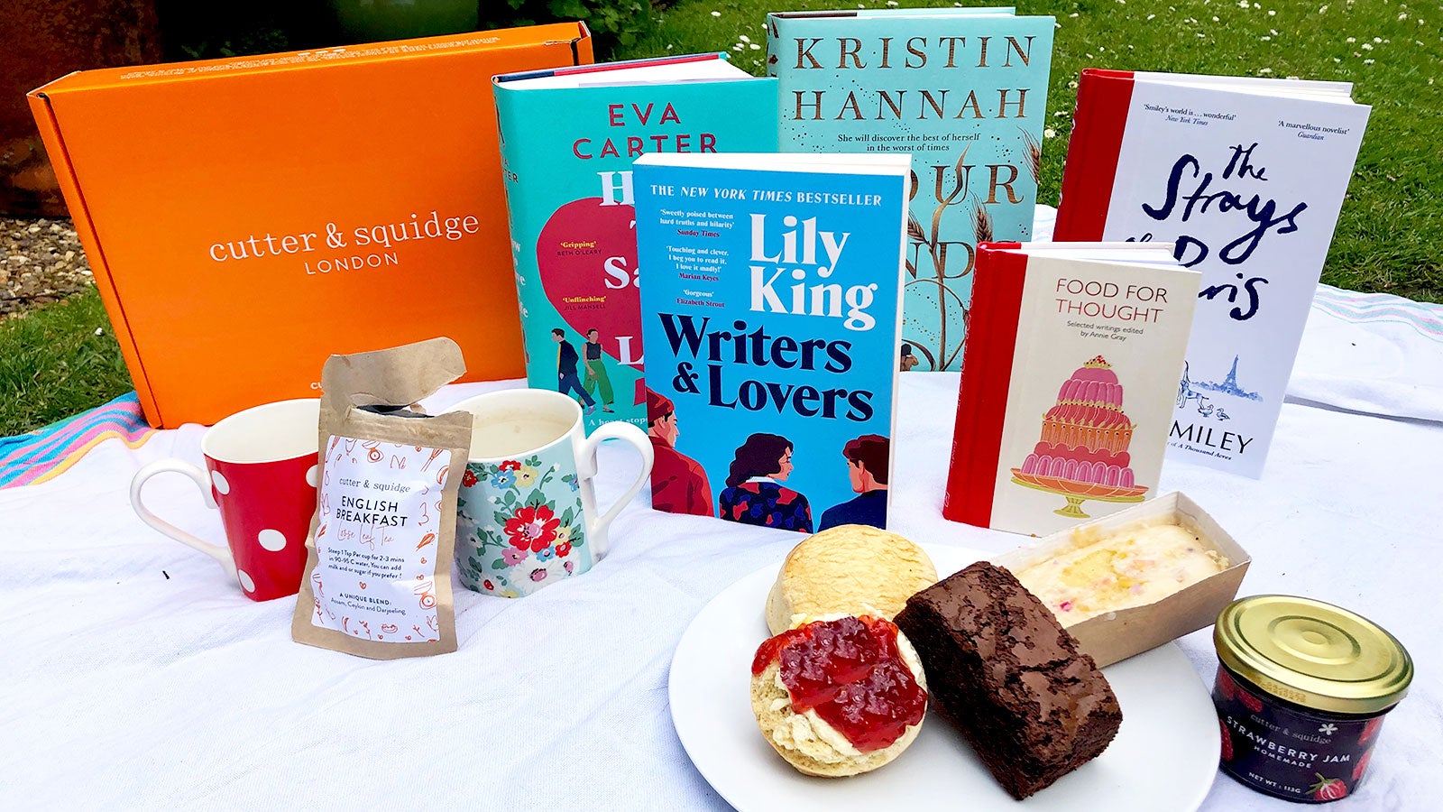 Five books stand propped on a white picnic blanket. They are surrounded by an afternoon tea with a Cutter & Squidge box.