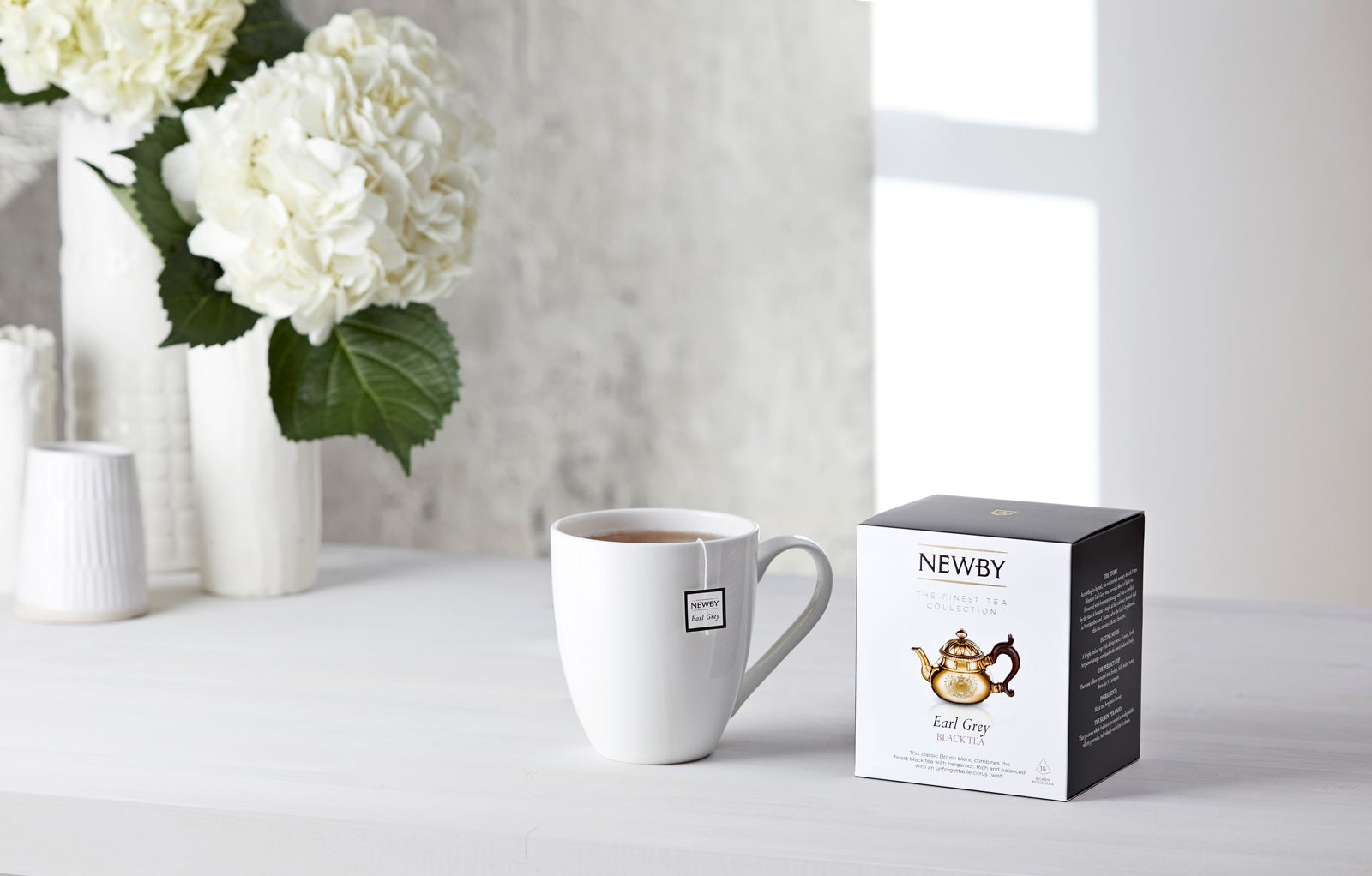 A white cup full of Earl Grey tea sits next to a box of tea, on a white table with white flowers in the background