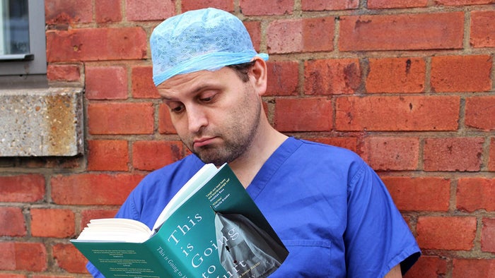 Adam Kay dressed in scrubs reading his own book