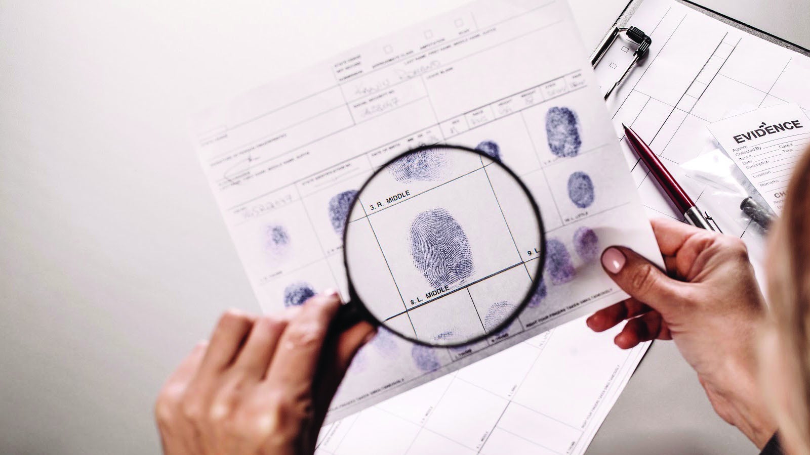 A pair of hands closely examines a set of fingerprints with a magnifying glass.