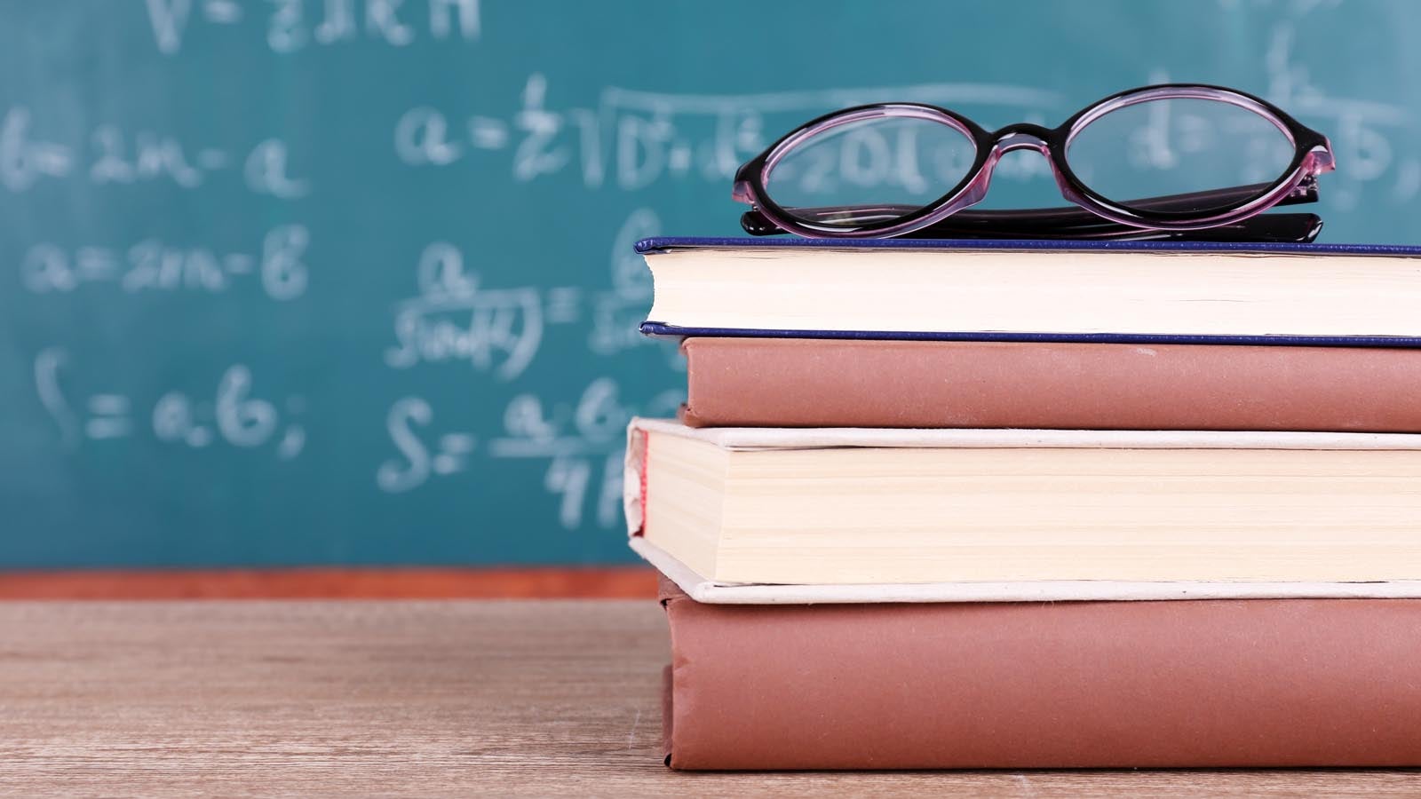 A pair of glasses on top of a stack of books, with a blurred chalkboard in the background