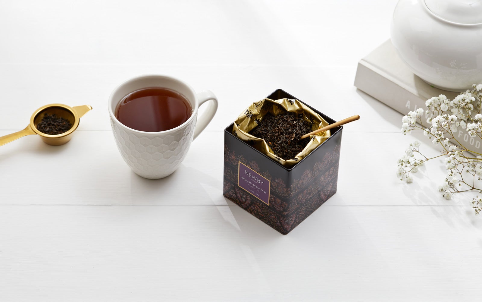 a box of loose leaf English Breakfast tea sits next to a white cup of tea and a gold strainer