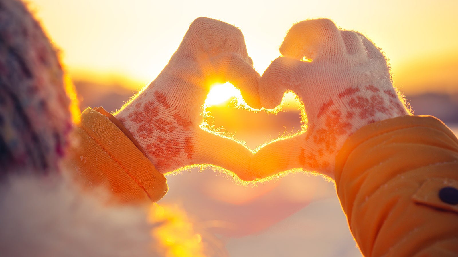 Two gloved hands making the shape of a heart in front of a winter sunset