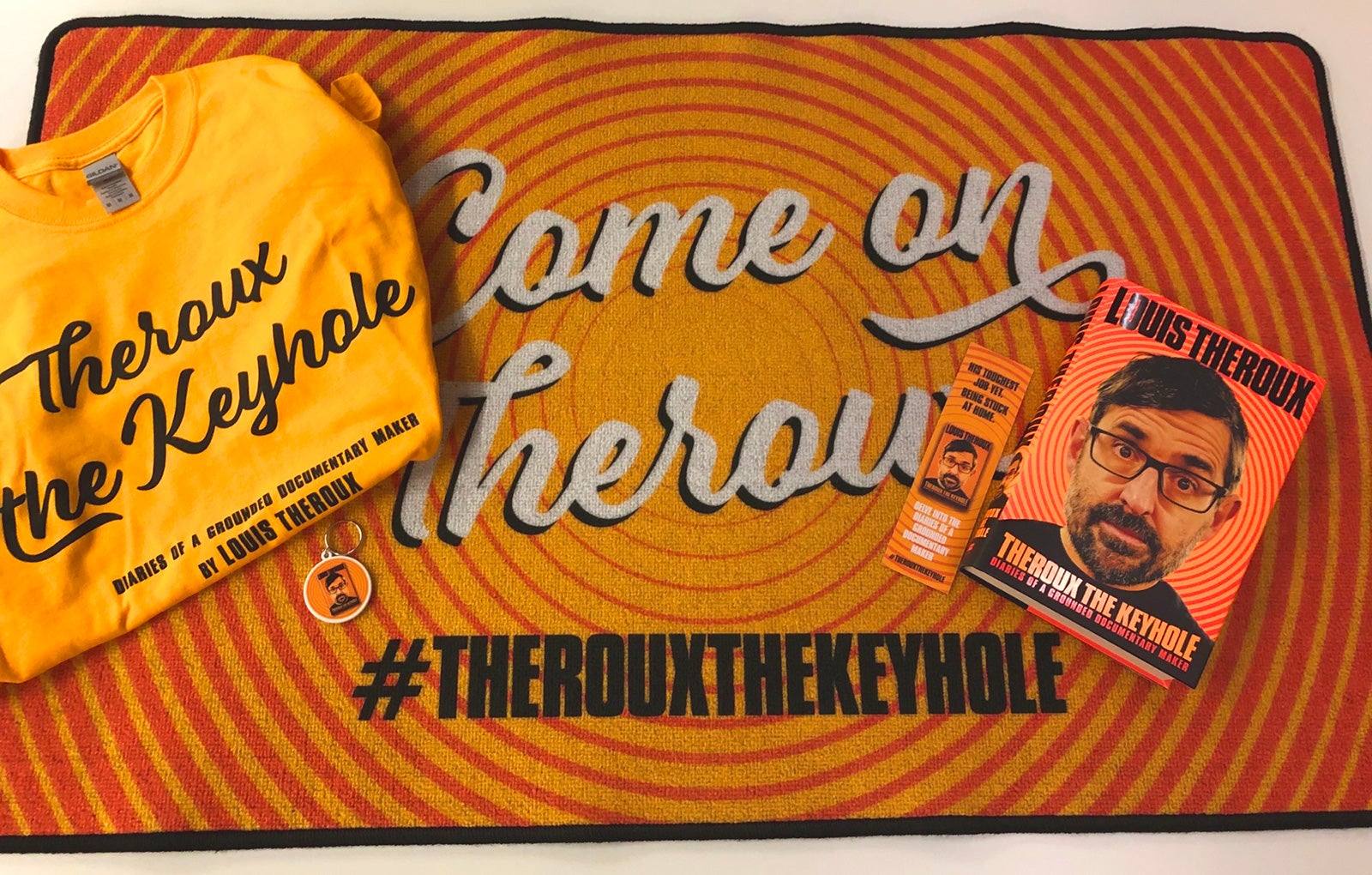 A copy of Theroux the Keyhole, a themed keyring, t-shirt and bookmark all sit atop a themed doormat.