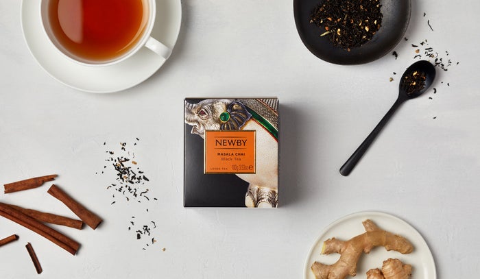 A box of Masala Chai tea sits at the centre of a white surface, surrounded by sprinklings of loose tea leaves, cinnamon sticks and ginger