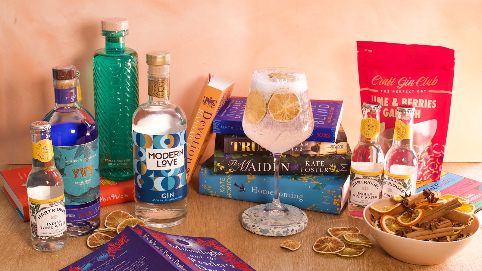An array of books displayed along with bottles of gin, a gin cocktail, some garnishes and tonics on a wooden table. 