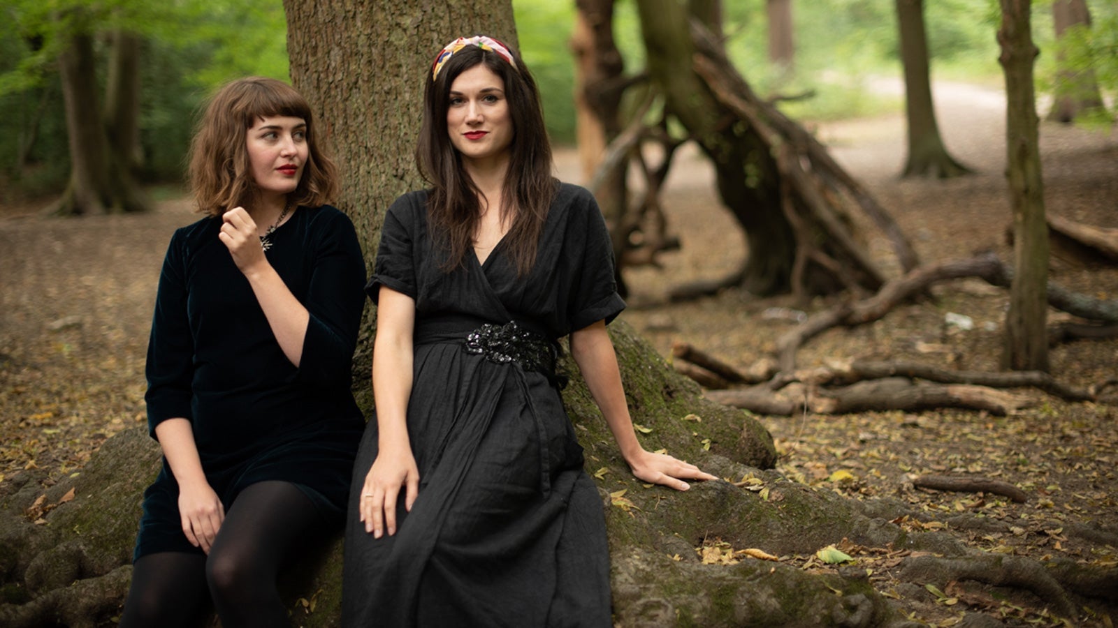 Jen Cownie and Fiona Lensvelt smiling, sit at the base of a tree in woodland