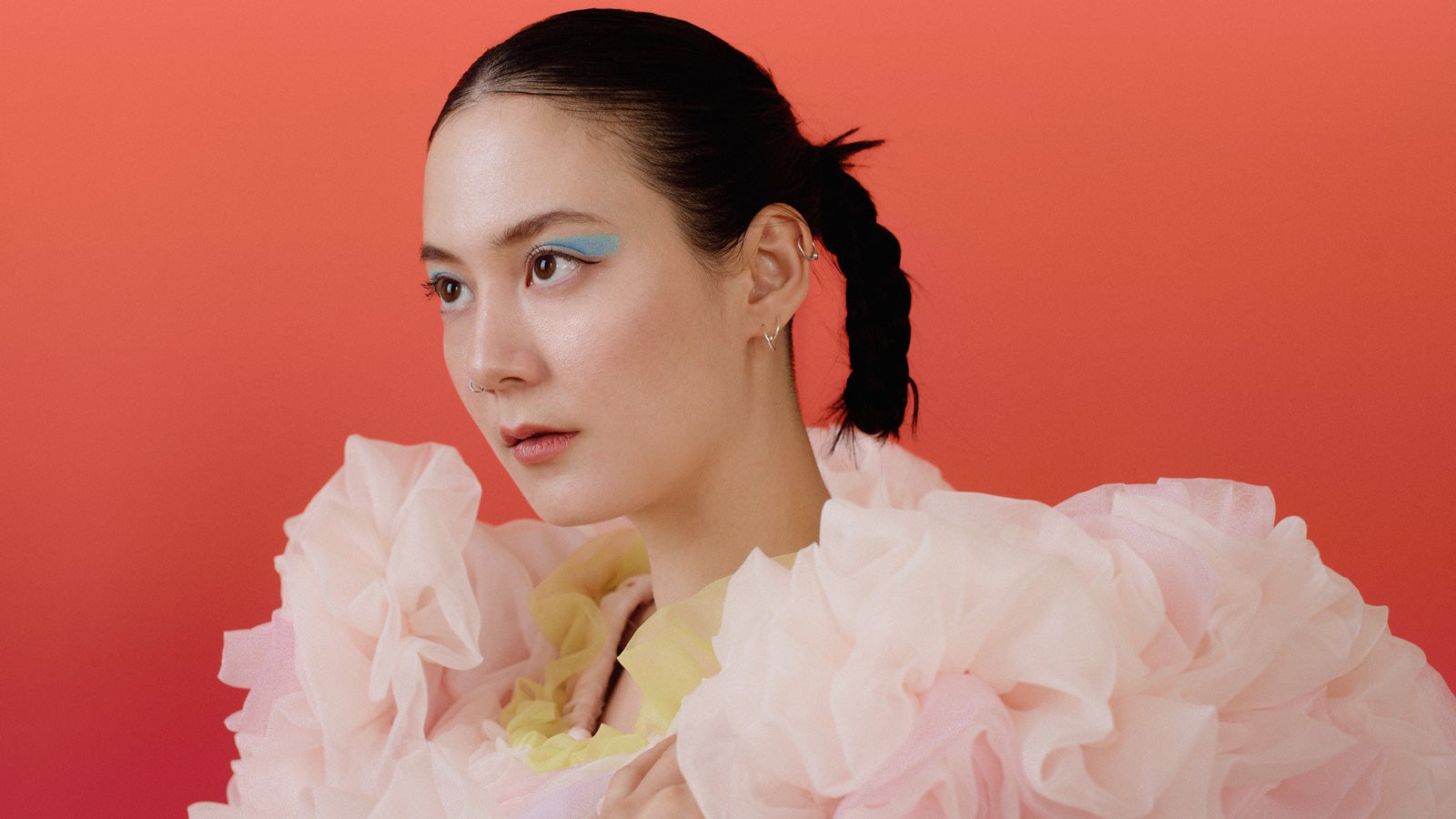 Michelle Zauner wears an oversized pink ruffled top and bright blue eye shadow, looking away from camera in front of a red/orange backdrop