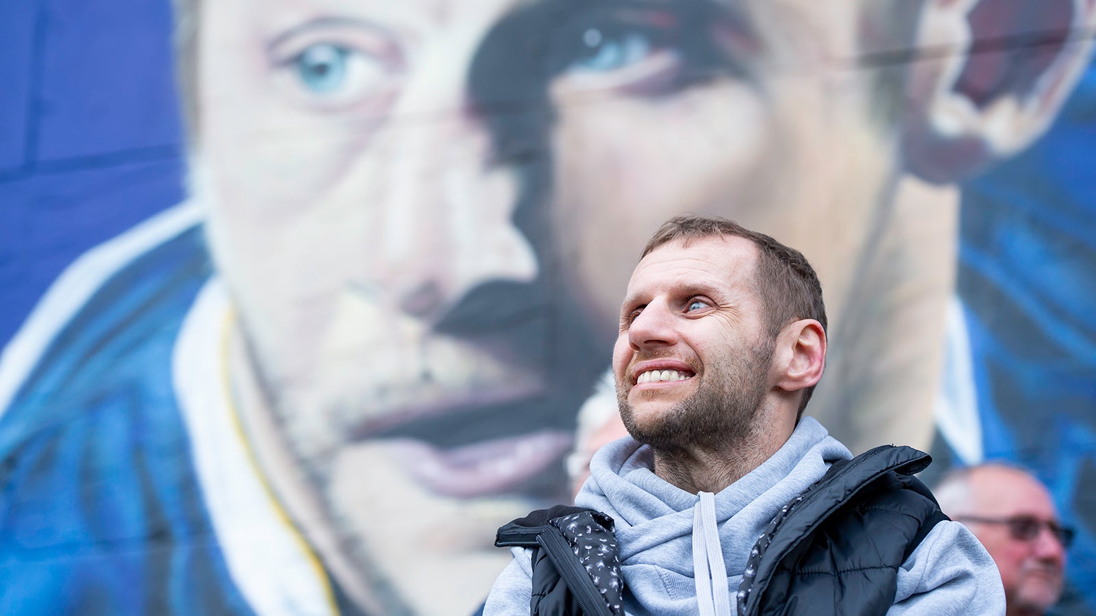 Rob Burrow smiles looking away front he camera in front o a mural of him playing rugby
