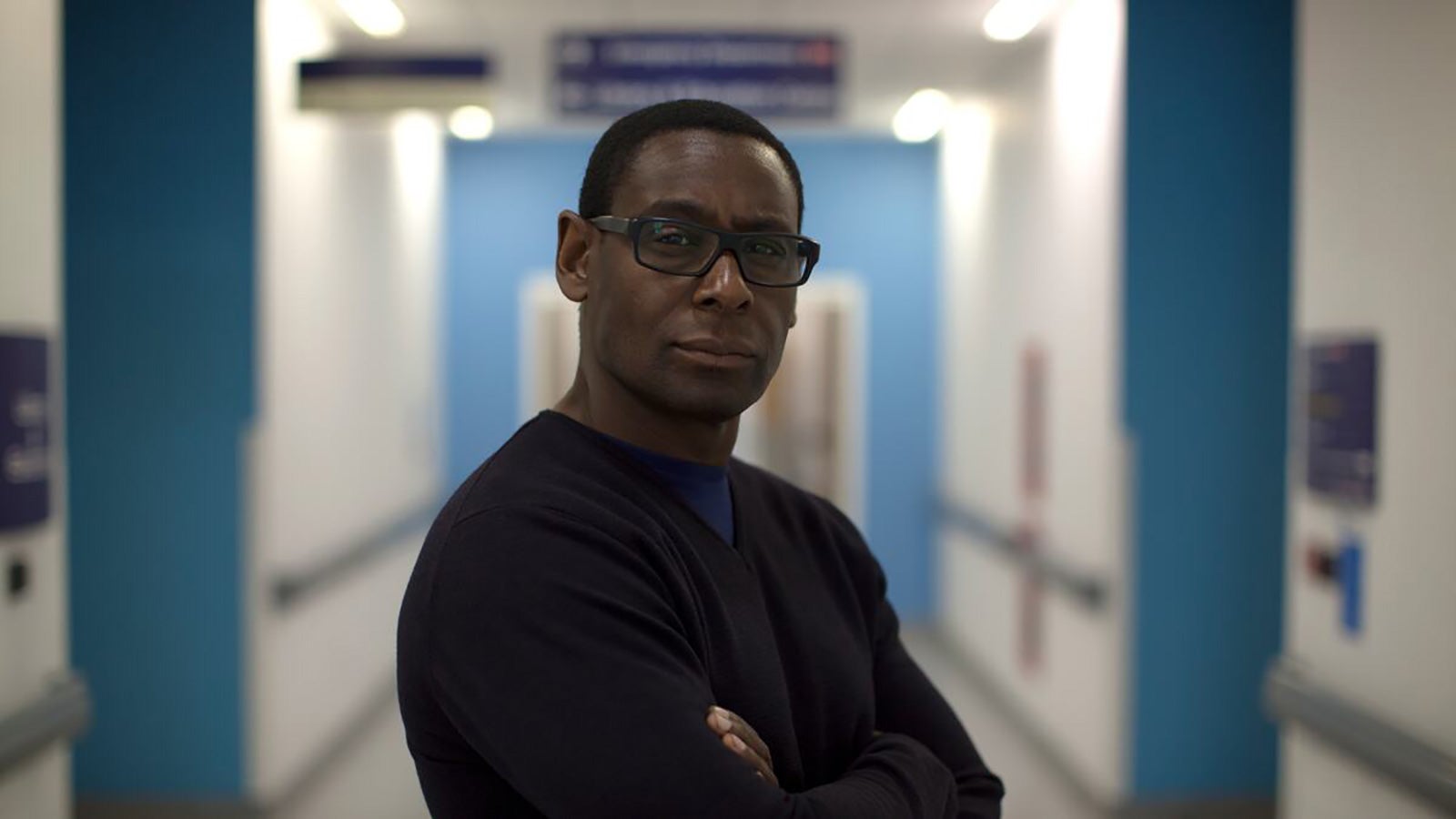 Actor David Harewood stands facing the camera with his arms crossed in a hospital ward.