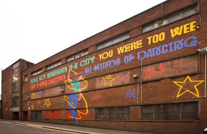 The side of Barrowland, an industrial looking red brick building, painted with a large 30m mural which shows a young boy spinning, surrounded by stars, and above it the words: 'You'll not remember the city. You were too wee. But there's dancing, all kinds of dancing'