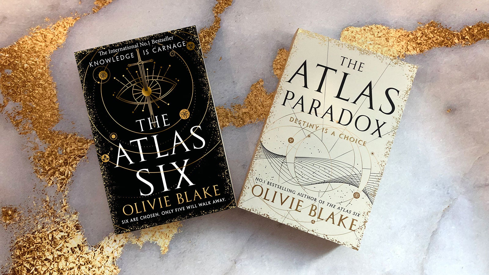 A paperback copy of The Atlas Six next to a proof copy of The Atlas Paradox on a white marble and gold background. 