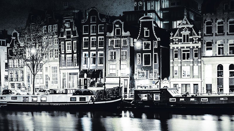 A black and white photograph of an Amsterdam street at night. Lights are on in some of the canal houses and a canal boat idles on the water. 