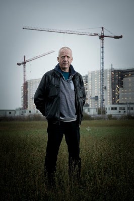 Image of author Neal Asher standing in a field with cranes and high rise buildings in the background