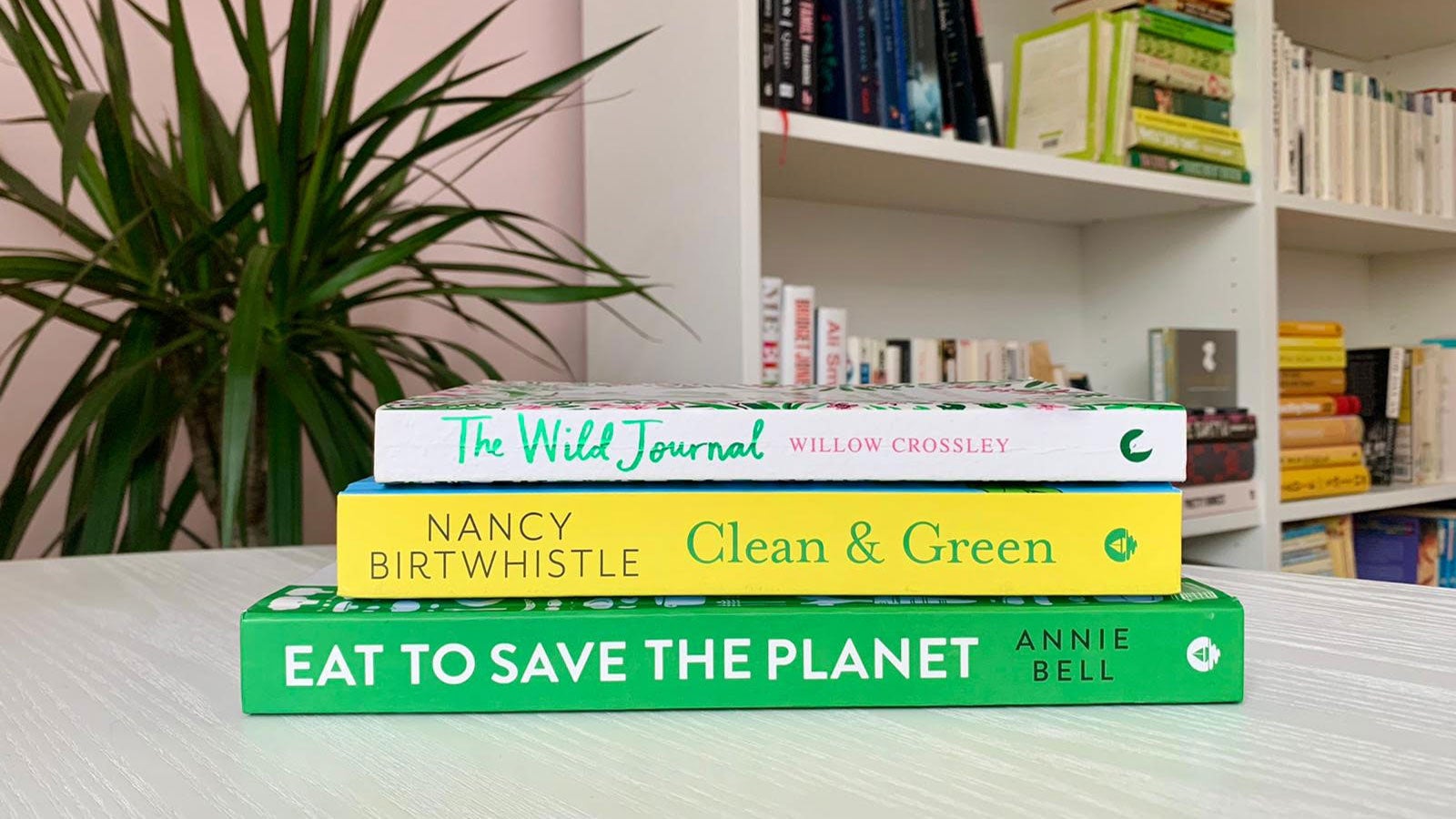 Eat to Save the Planet, Clean & Green, The Wild Journal stacked on a white table in front of a bookcase and a plant.